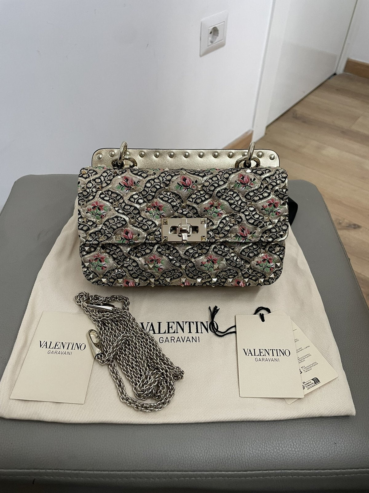 Borsa a spalla Valentino Garavani Rockstud Spike piccola poggiata su pouf grigio con la sua dustbag originale e tracolla a catena metallica inclusa.